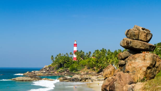 The lighthouse on the cape stretching into the Arabian Sea and beach with rocks in the foreground at a resort in Kerala in sunny weather (southern India)