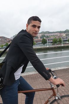 Close up or a young man riding on a bike on a cloudy day near the river with a shoulder bag and jeans on a cloudy day