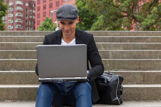 Young man with Irish beret sitting on a wall next to an urban stairs and a shoulder bag near