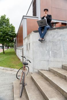 Young man with Irish beret sitting on a wall with a laptop next to a bike