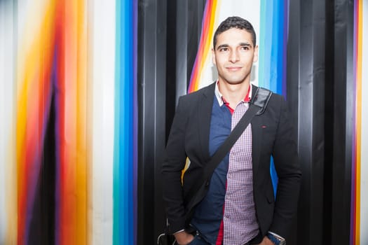 Modern young man with Arab features with shoulder bag and jacket without a tie on multicolored background and the hands in his pocket horizontal photography