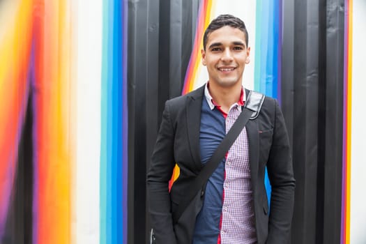 Modern young man with Arab features with shoulder bag and jacket without a tie on multicolored background and the hands in his pocket horizontal photography