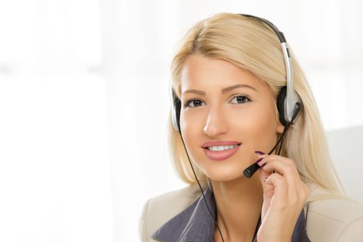 Close-up of a portrait young beautiful blonde girl, elegantly dressed, sitting at an office with a headset on her head. With a smile looking at the camera.