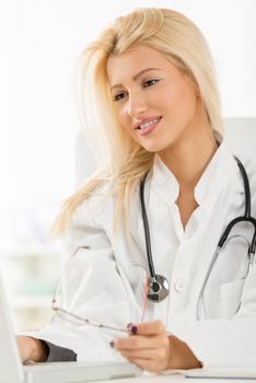 Attractive blonde female doctor in her consulting room, sitting at the table in front of laptop.