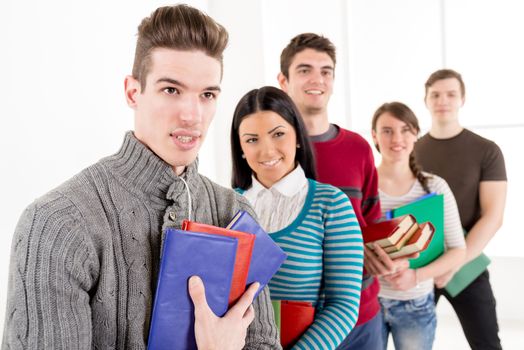 A beautiful smiling young man with book is standing in the foreground. A happy group of his friends standing In A Row is behind him. 