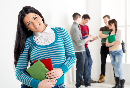 A beautiful smiling girl with book's is standing in the foreground. A group of her friends is behind her. Looking at camera.