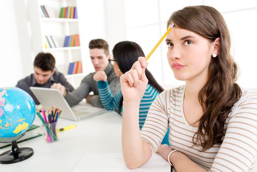 A beautiful smiling girl learning. She is sitting and thinking in the foreground. A happy group of her friends is behind him and looking at laptop. 