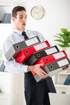 Young stressed Businessman standing in the office and holding many binders. They're spilling out hand.