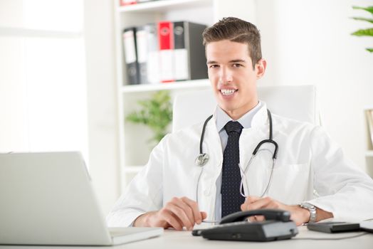 Young male doctor with stethoscope sitting at desk in doctor's room and looking at camera.