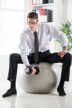 The young businessman sitting in the office on pilates ball and doing exercise with dumbbells.