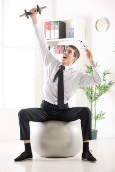 Successful businessman sitting in the office on pilates ball and doing exercise with dumbbells.