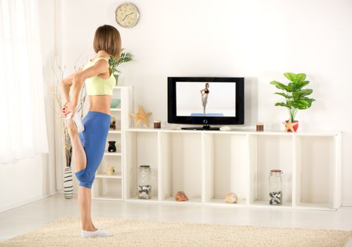 Young woman in sports clothes, doing stretching exercises in the room, in front TV.