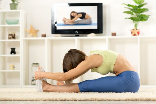 Young woman in sports clothes, doing stretching exercises in the room, in front TV.