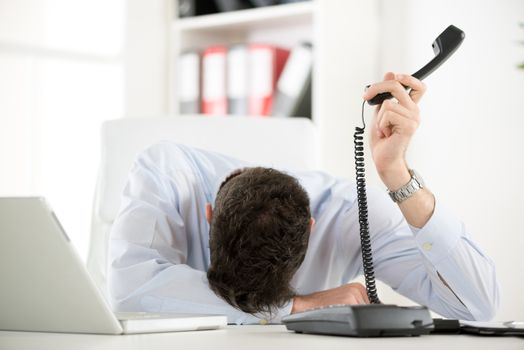 A young tired businesswoman, exhausted from work sleeping in front of his laptop, leaning on office desk with a telephone handset on hand.