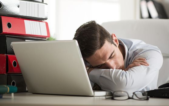 Young tired Businessman sleeping in the office. In front of his a laptop and bunch of binders.