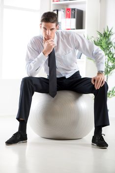 The young businessman sitting in the office on pilates ball and thinking. Looking at camera.