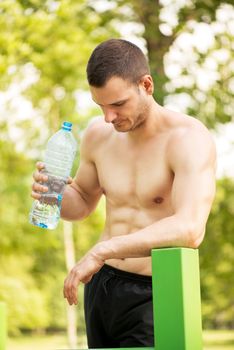 Naked muscle man resting after exercising in the outdoor gym. He is holding water bottle and thinking.
