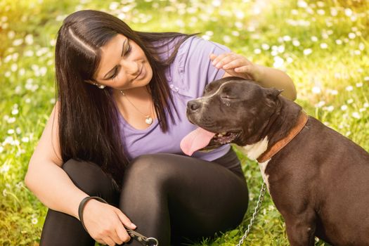 Beautiful young woman with her cute stafford terrier sitting on grass in the park and enjoying.