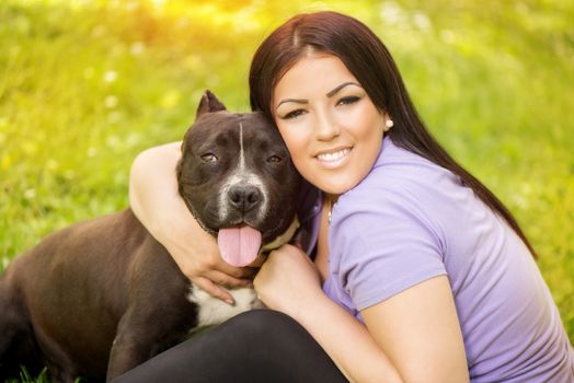 Beautiful young woman hugging her cute stafford terrier. They sitting on grass in the park and enjoying. Looking at cemera.