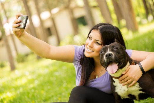 Beautiful young woman hugging her cute stafford terrier. They sitting on grass in the park and taking Self portrait on a smartphone. 