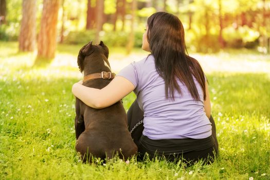 Beautiful young woman hugging her cute stafford terrier. They sitting on grass in the park and enjoying. Rear view.