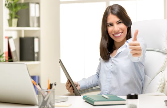 Successful businesswoman sitting at the desk in office with thumb up. Looking at camera and smiling.