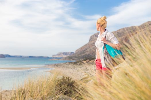 Relaxed woman on vacations in white loose shirt carrying beach bag and towel, enjoying beautiful coastline view of white sandy lagoon at Balos beach, Greece.