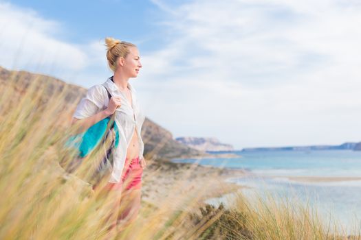 Relaxed woman on vacations in white loose shirt carrying beach bag and towel, enjoying beautiful coastline view of white sandy lagoon at Balos beach, Greece.