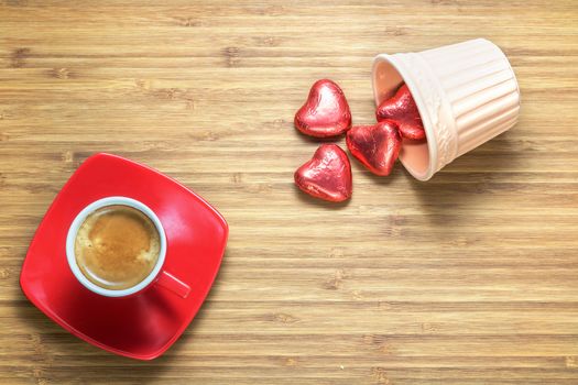 Heart shaped sweets wrapped in a bright red foil lying on a wooden texture with red cup of coffe near it. Background for romantic themes.