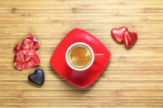 Heart shaped sweets wrapped in a bright red foil lying on a wooden texture with red cup of coffe near it. Background for romantic themes.