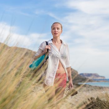 Relaxed woman on vacations in white loose shirt carrying beach bag and towel, enjoying beautiful coastline view of white sandy lagoon at Balos beach, Greece.