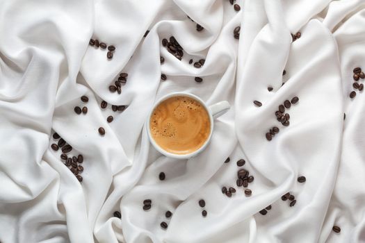 Cup of coffee on a white silk fabric. Espresso with froth in the form of smiley face and scattered beans. Top view