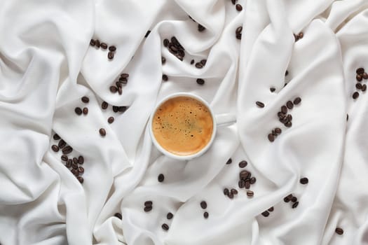 Cup of coffee on a white silk fabric. Espresso and scattered beans. Top view