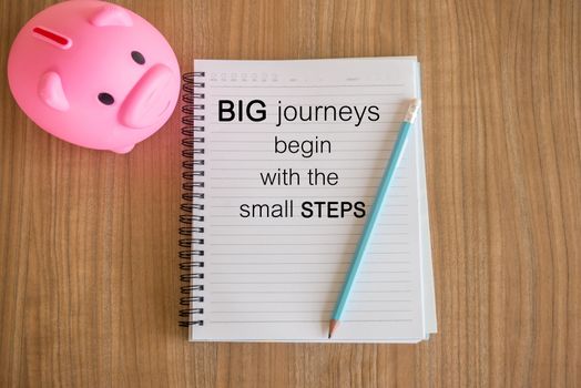 Word  Big journeys begin with the small steps.Inspirational motivational quote on paper and a piggy bank on the wooden table background
