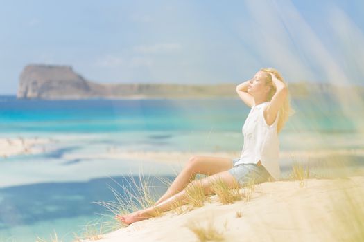 Relaxed woman enjoying sun, freedom and life an a beautiful sandy beach of Balos in Greece. Young lady feeling free, relaxed and happy. Vacations, freedom, happiness, enjoyment and well being.