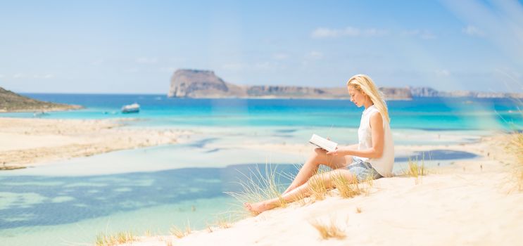 Relaxed woman enjoying sun, freedom and good book an beautiful sandy beach of Balos in Greece. Young lady reading, feeling free and relaxed. Vacations, freedom, happiness, enjoyment and well being.