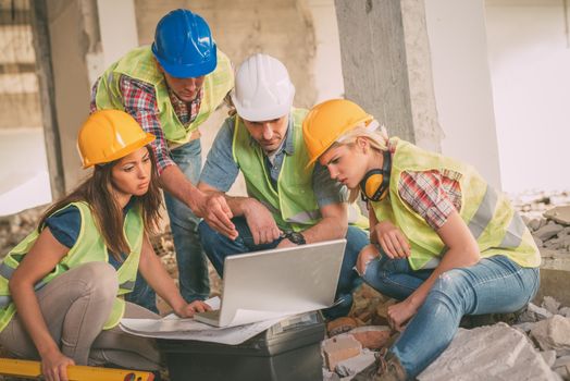 Four construction architects review plan at laptop in front building damaged in the disaster.