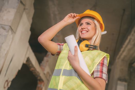 Beautiful young female construction architects at a construction site. She is holding plans and smiling holding helmet.
