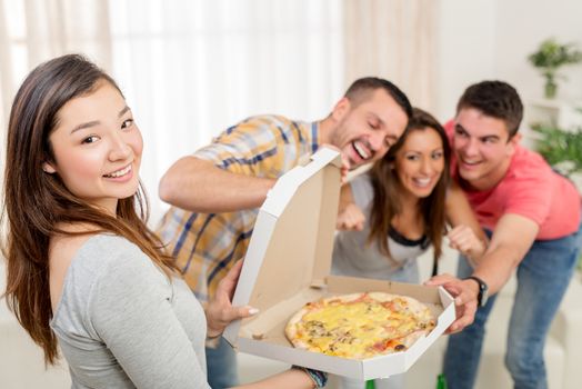 Four cheerful friends enjoying pizza together at home party. Selective focus. Focus on foreground, on Japanese girl. She is holding box with pizza and looking at camera.