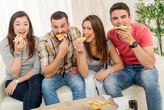 Four cheerful friends hanging out in an apartment. They eating pizza and looking at camera.