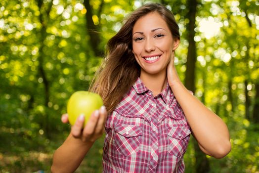 Beautiful smiling girl in the nature holding green apple and looking at camera.