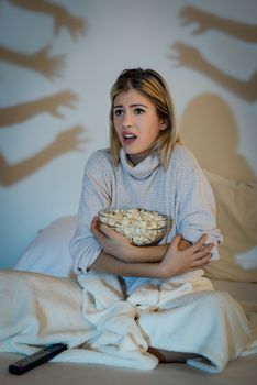 Beautiful young woman watching a horror movie in the bed. She holding popcorn and looking tv with fear on her face.