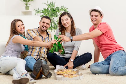 Four cheerful friends hanging out in an apartment. They drinking beer and eating pizza. Looking at camera.