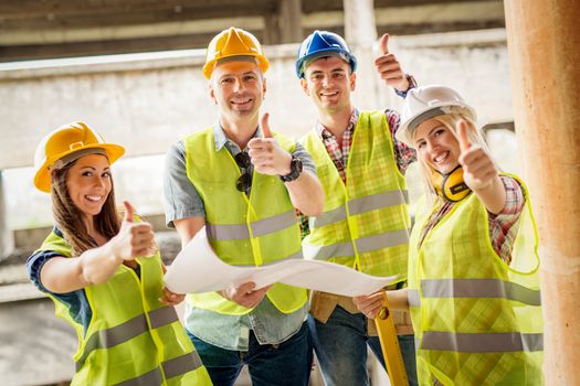 Four construction architects review plan in front building damaged in the disaster. They are showing thumbs up and looking at the camera.