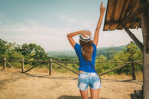 Cheerful woman on summer travel vacation standing by viewpoint with outstretched arm. Rear view.
