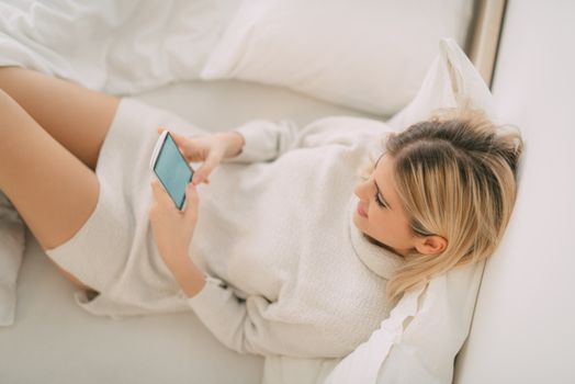Beautiful young smiling woman relaxing in bed and typing message on smart phone.