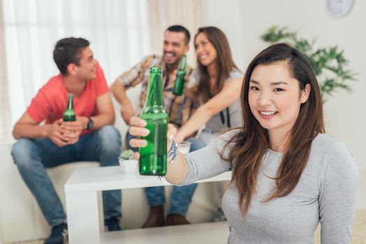 Close up of a young Japanese girl smiling at home party with beer and cheers. Her friends in the background. Selectiv focus. Focus on foreground.