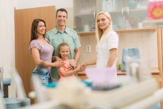 Little girl and her parents visiting dentist. The little girl is shaking hand with female dentist and smiling. Looking at camera.