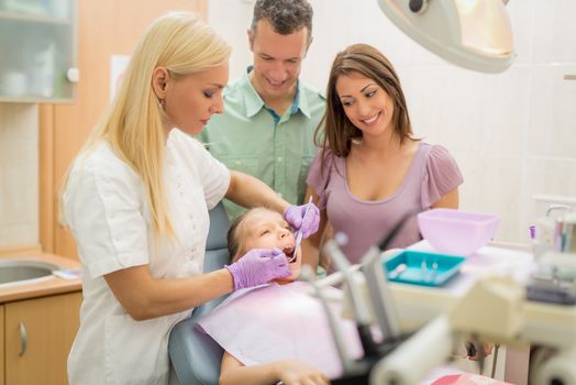 Happy family at visit in the dentist office. Female dentist checking teeth the little girl, her parents standing next to her.