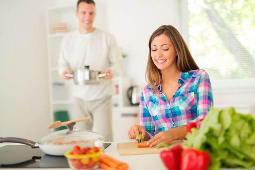 Beautiful young woman cooking healthy meal in the domestic kitchen. Her boyfriend behind, holding pot. Selective focus. Focus on foreground.